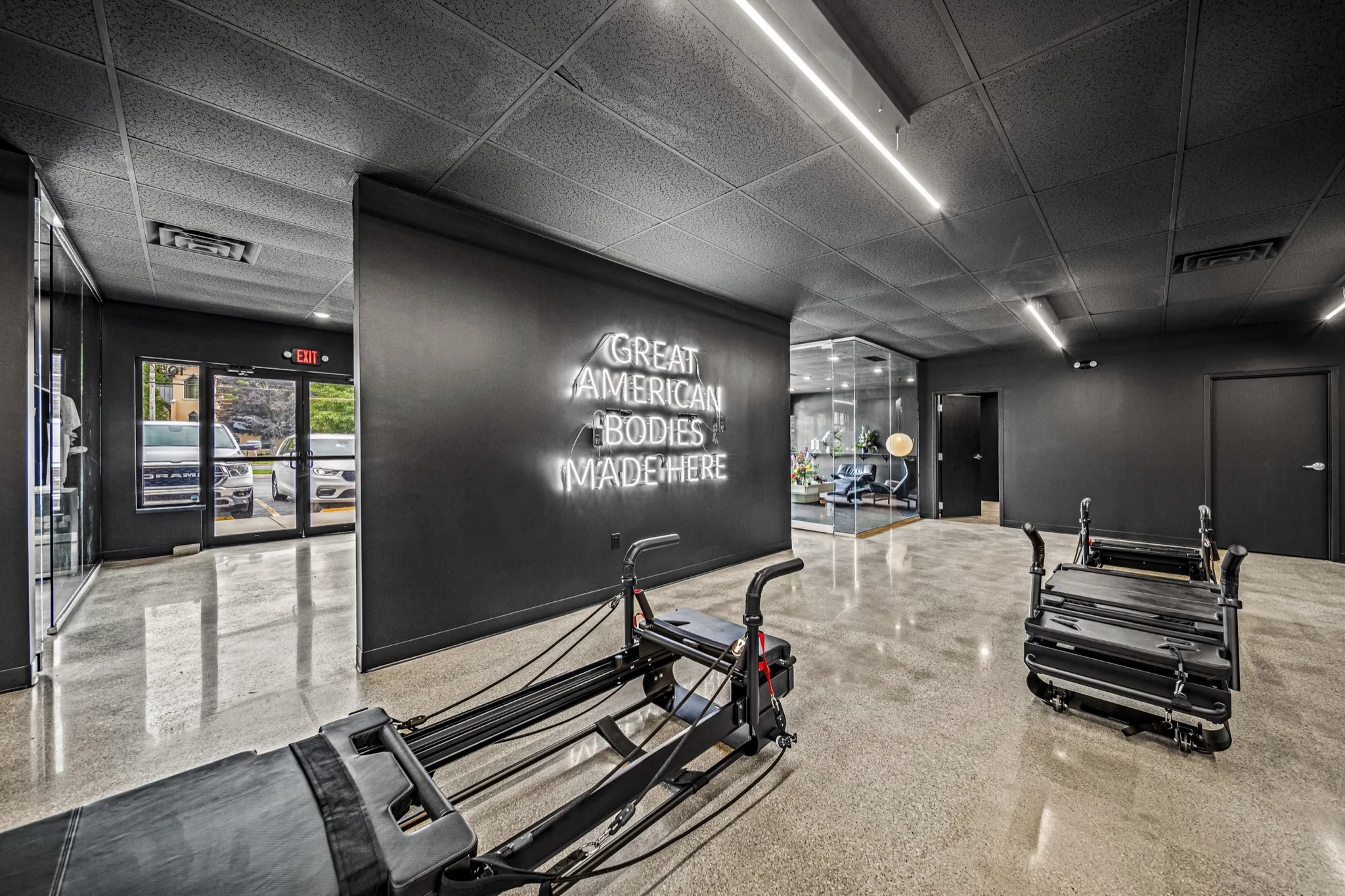 Neon Great American Bodies Made Here sign with Pilates reformer in foreground, polished concrete — commercial fitness interior by Skoda Construction, Northeast Ohio