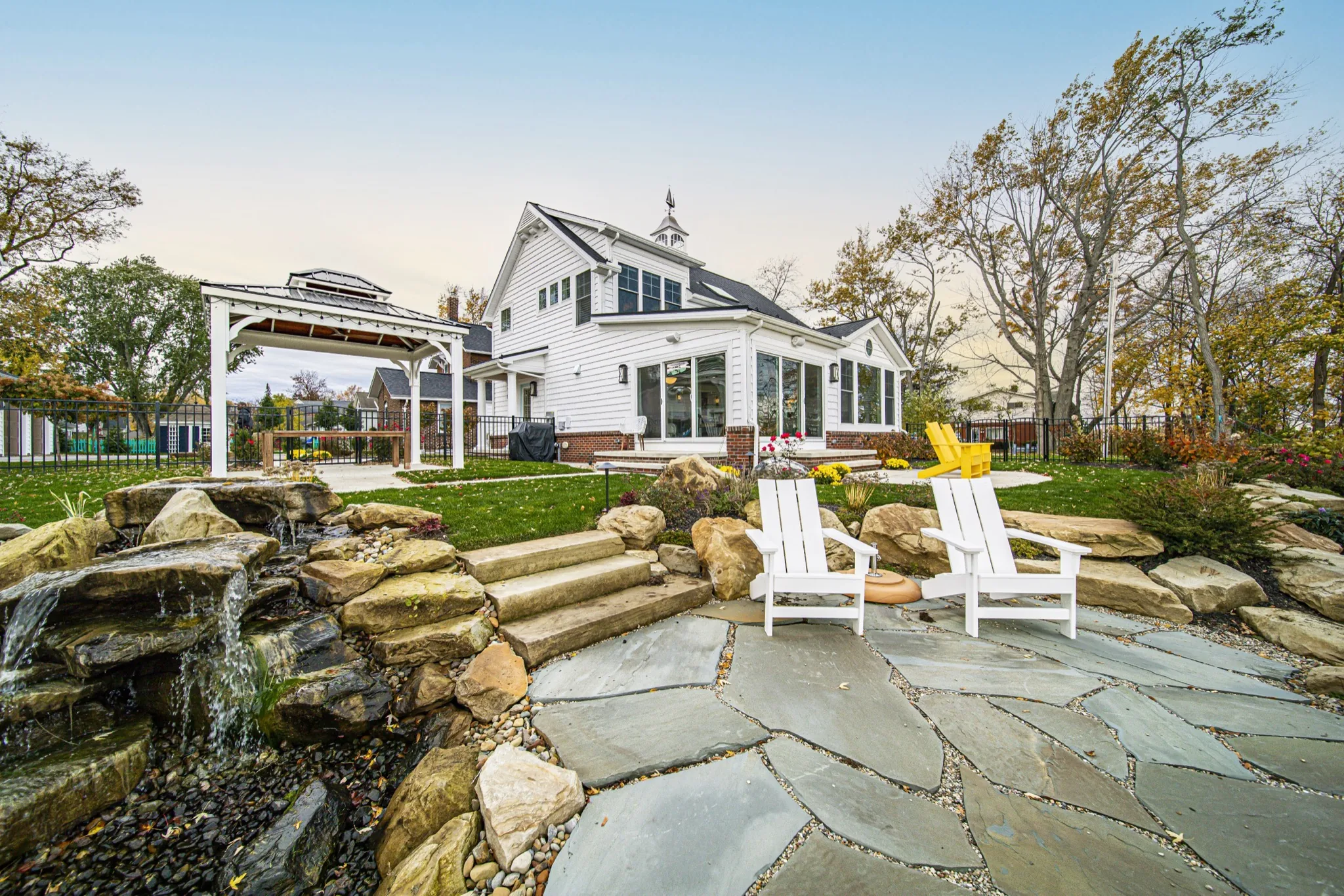 Stone waterfall patio with gazebo and white farmhouse at twilight — custom outdoor living by Skoda Construction, Northeast Ohio