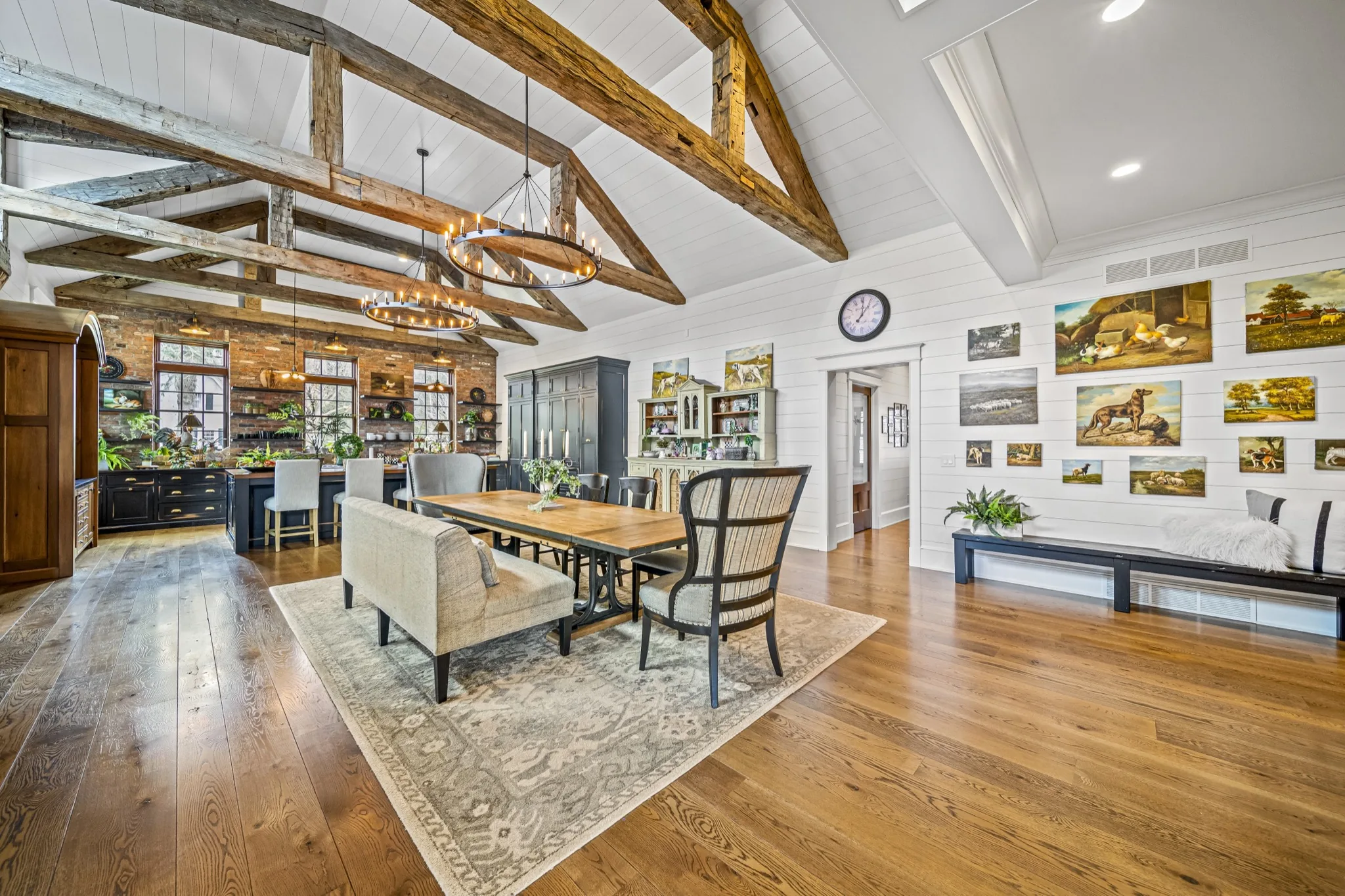 Panoramic view of open great room, dining, and kitchen with reclaimed timber trusses, iron chandelier, shiplap gallery wall, and exposed brick — luxury farmhouse by Skoda Construction, Northeast Ohio