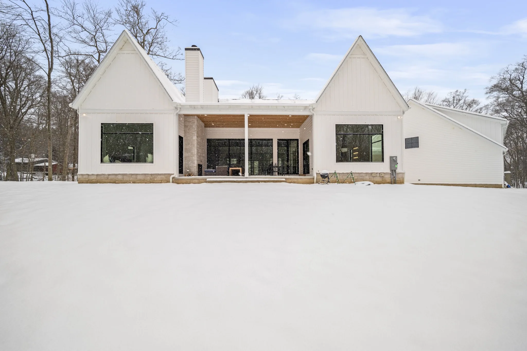 Rear elevation of modern luxury estate featuring covered porch, natural stone chimney, and walls of glass overlooking a private wooded lot blanketed in snow — Skoda Construction