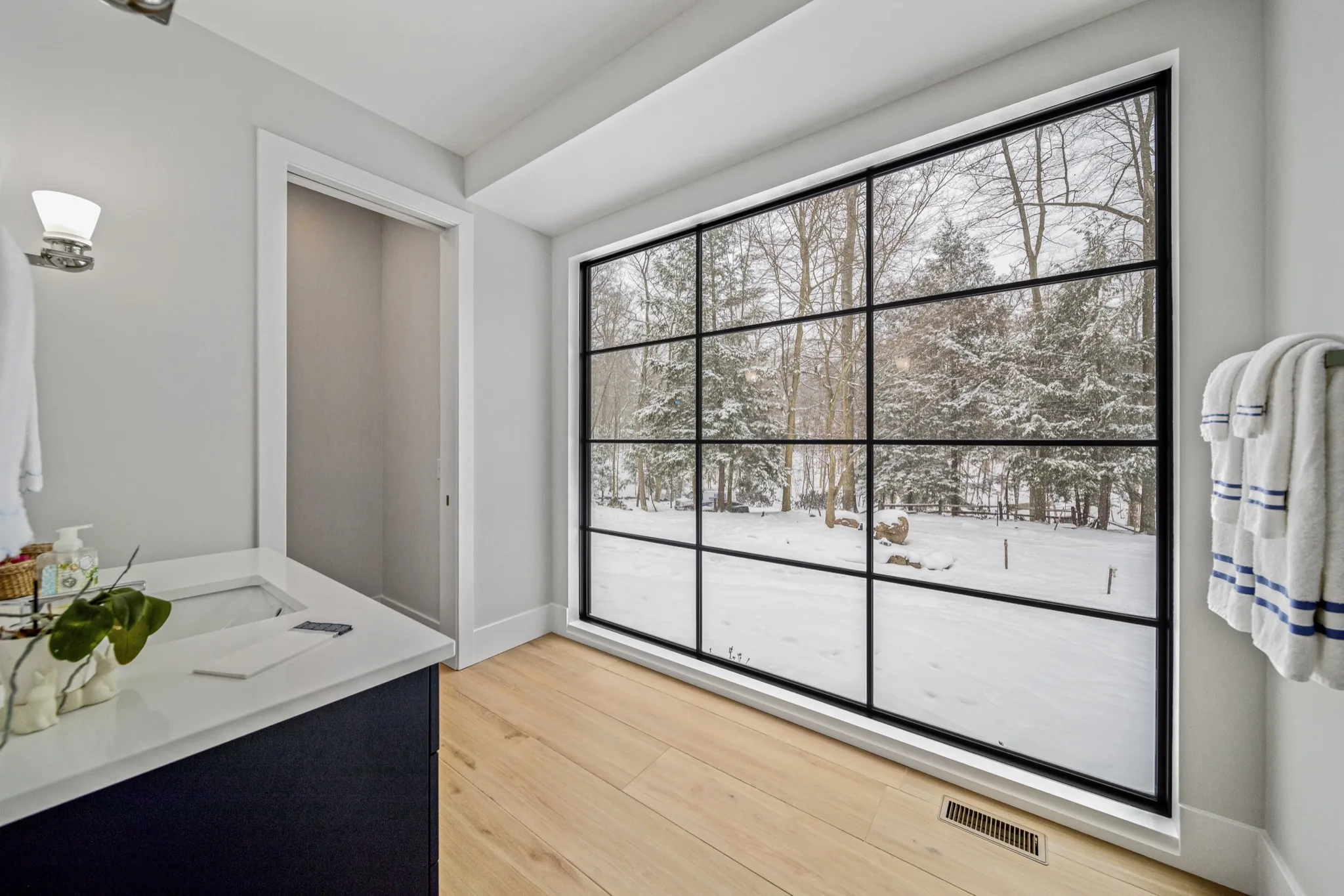 Bathroom with floor-to-ceiling grid-paned window overlooking snow-covered woodland, dark vanity, and quartz countertop — modern luxury home by Skoda Construction, Northeast Ohio