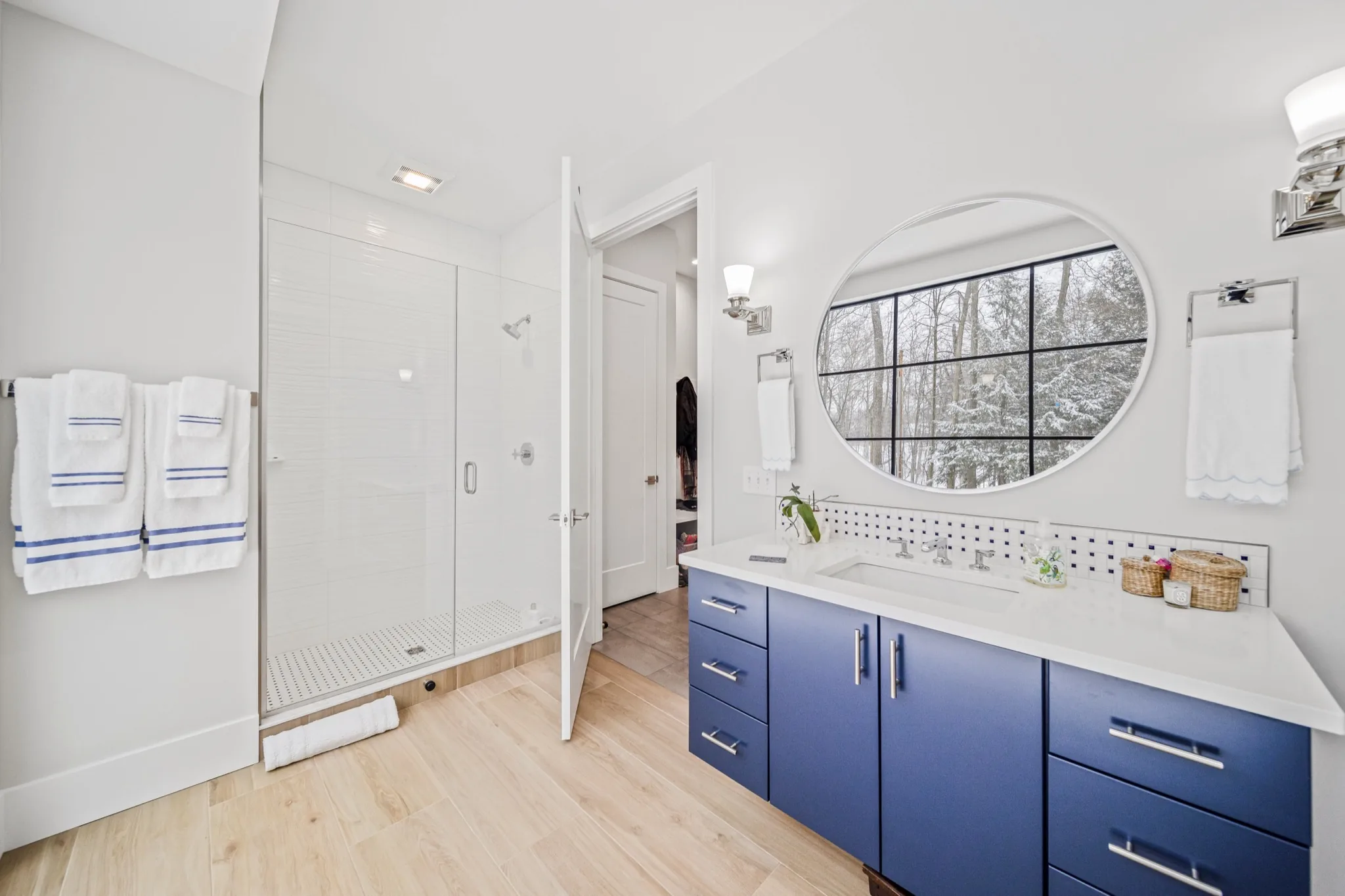 Guest bathroom with bold blue floating vanity, oversized round porthole window overlooking snowy woods, and glass shower enclosure — luxury home by Skoda Construction, Northeast Ohio