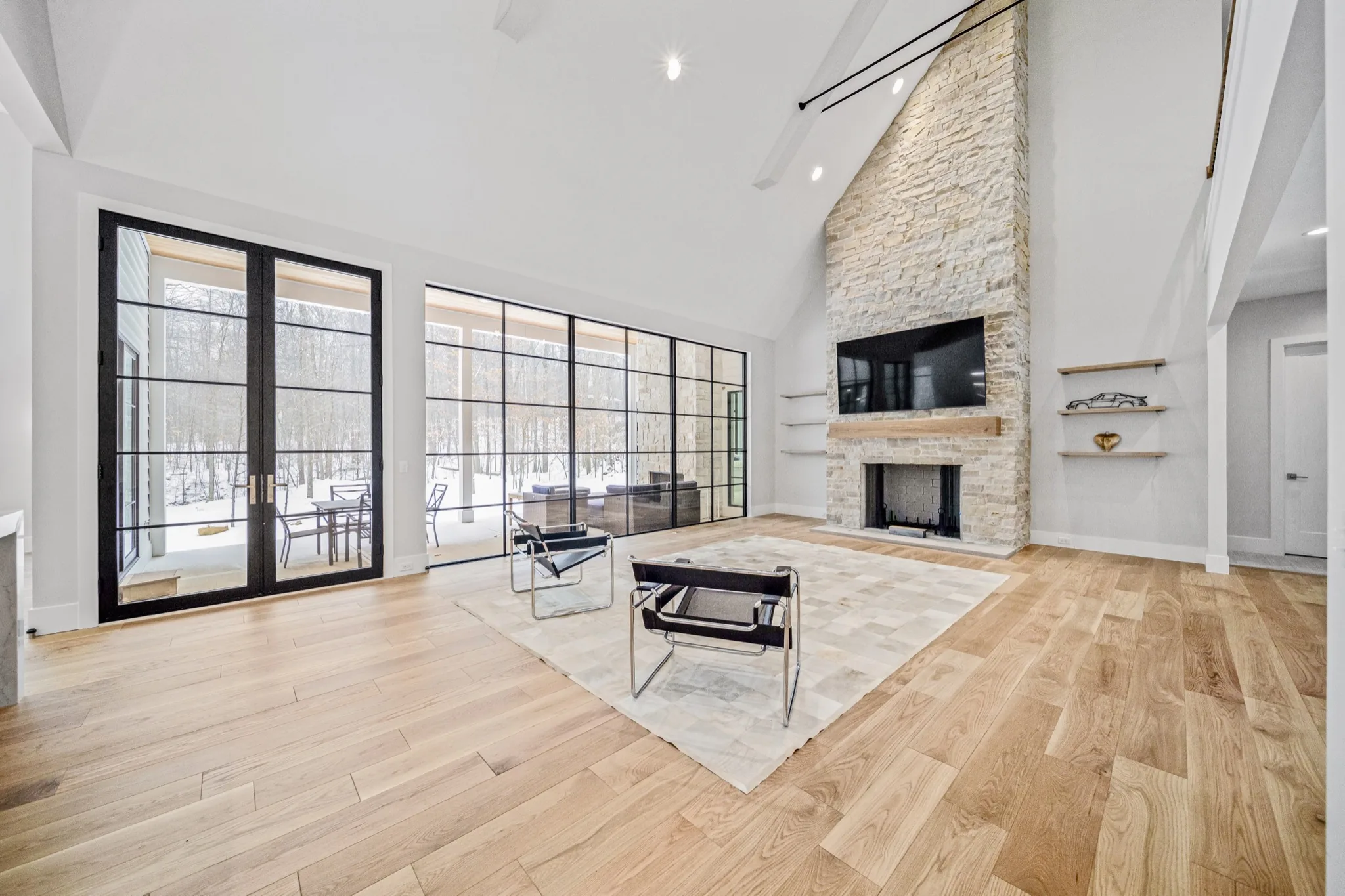 Modern great room with floor-to-ceiling glass wall revealing snow-covered woodland, natural stone fireplace, and covered outdoor living space beyond — Skoda Construction luxury estate