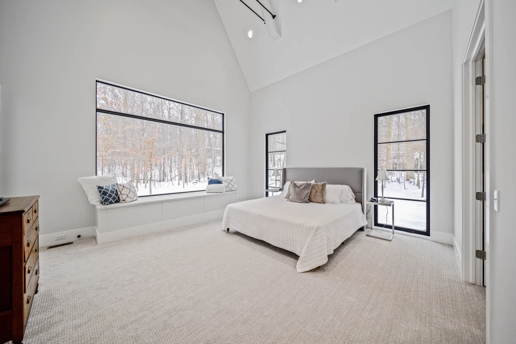 Luxurious primary bedroom with vaulted ceiling, oversized picture window with built-in window seat, and floor-to-ceiling glass door overlooking a private wooded setting — Skoda Construction custom home