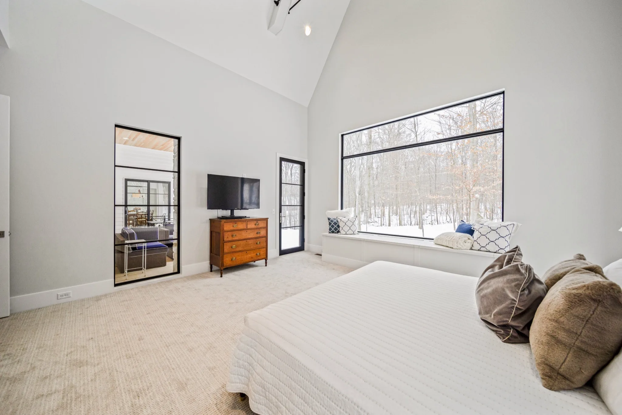 Primary bedroom with vaulted ceiling, oversized picture window with built-in window seat, and glass door to private balcony overlooking snowy woodland — Skoda Construction luxury estate