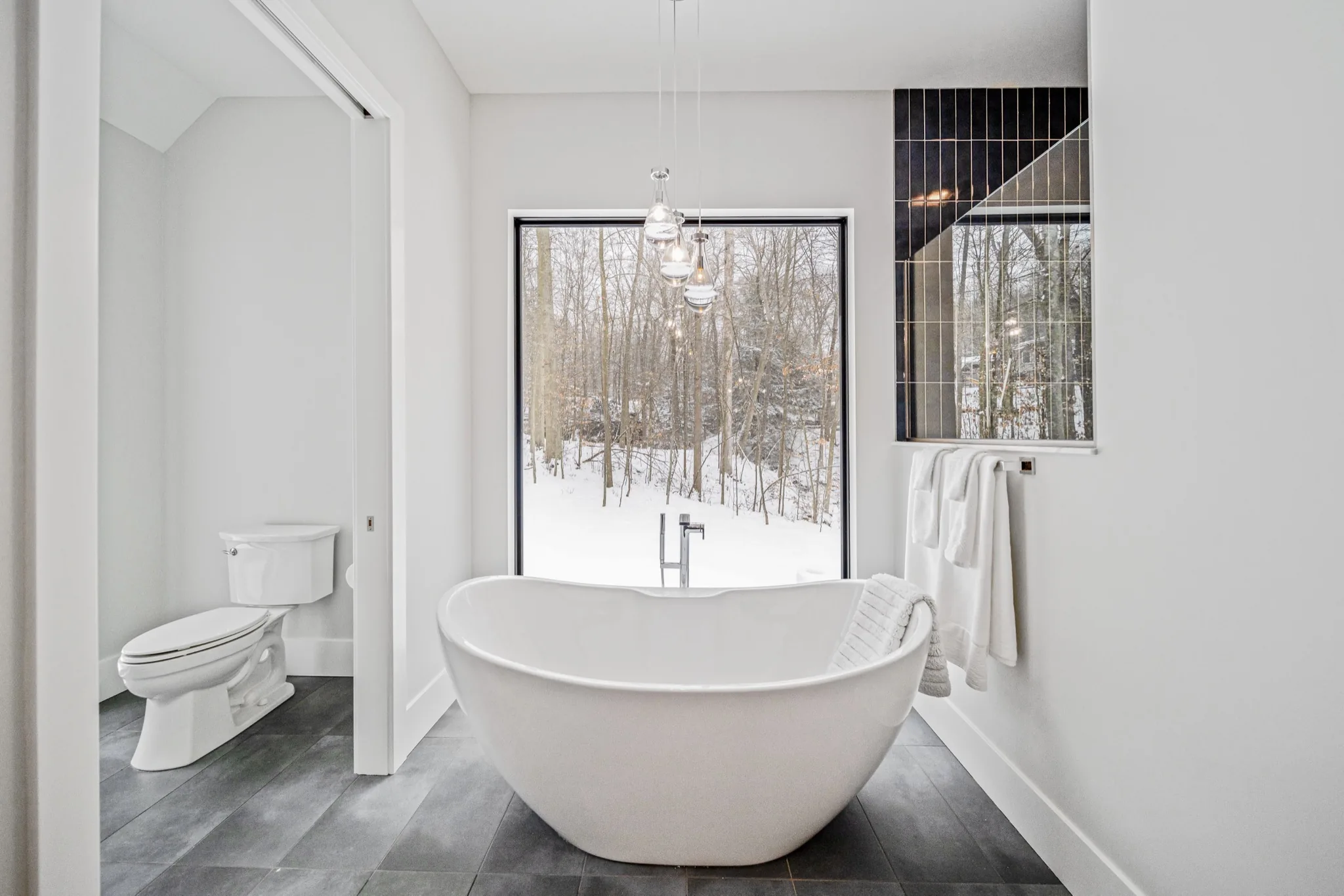 Freestanding soaking tub framed by a floor-to-ceiling picture window overlooking snow-covered woods with crystal pendant lighting — luxury primary bathroom by Skoda Construction