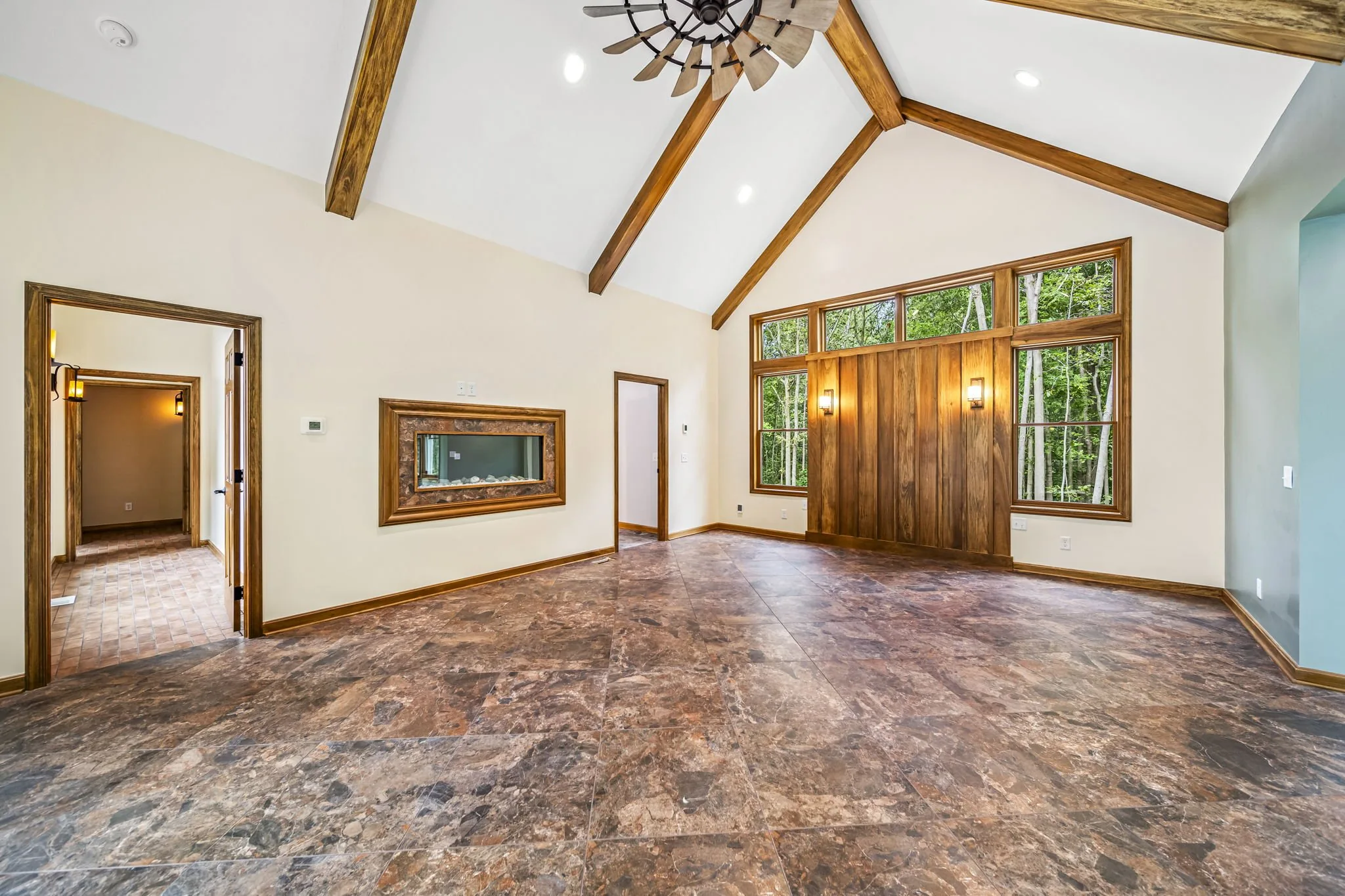 Vaulted great room with exposed timber beams, windmill ceiling fan, floor-to-ceiling windows overlooking forest, and natural stone accents — custom estate by Skoda Construction, Northeast Ohio