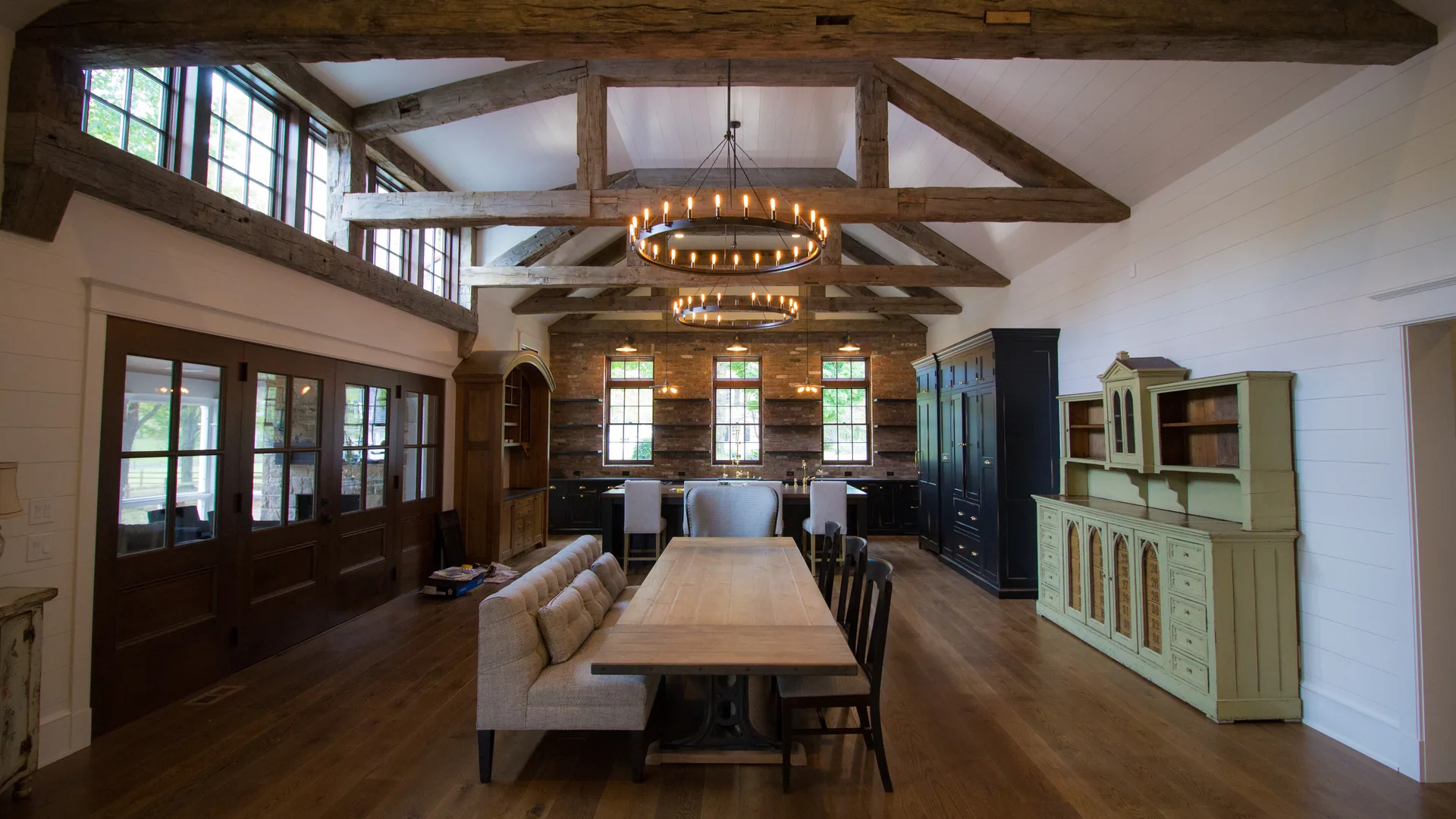 Two-story great room with reclaimed timber trusses, iron ring chandelier, exposed stone and reclaimed wood walls, farmhouse dining table — luxury custom home by Skoda Construction, Northeast Ohio